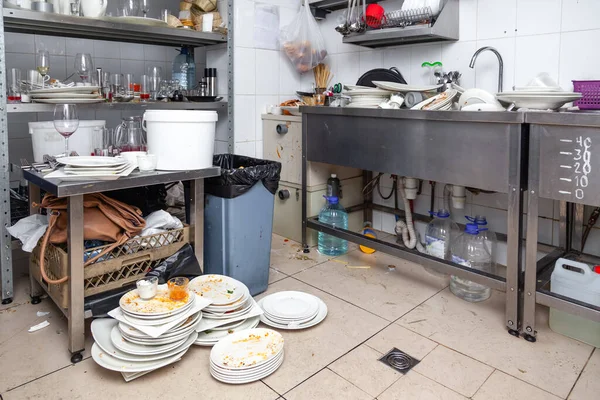 Cozinha de restaurante com louça acumulada, lixo no chão e bancada desorganizada durante o serviço
